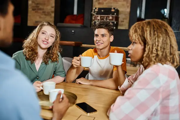 Amigos multiétnicos positivos con tazas de café hablando en la cafetería salón de los estudiantes albergue, viajeros - foto de stock