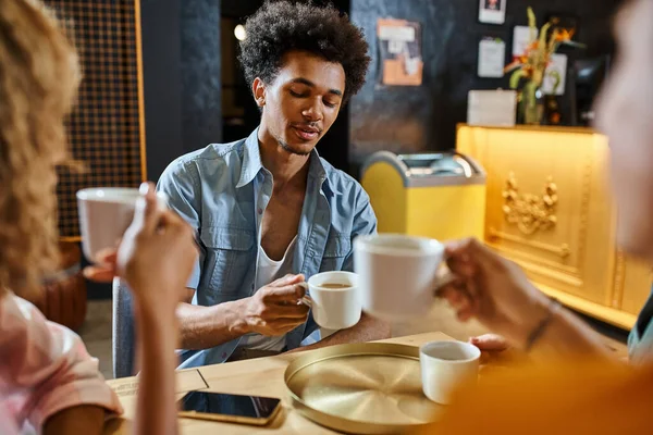 Young african american man sitting with coffee cup near multiethnic buddies in lobby cafe of hostel — Stock Photo