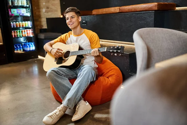 Joyeux jeune homme assis dans une chaise de sac de haricot et jouer de la guitare dans le hall de l'auberge d'étudiants modernes — Photo de stock