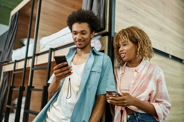 Sonriente afroamericano hombre navegar por Internet en el teléfono inteligente cerca de la novia en los estudiantes albergue - foto de stock