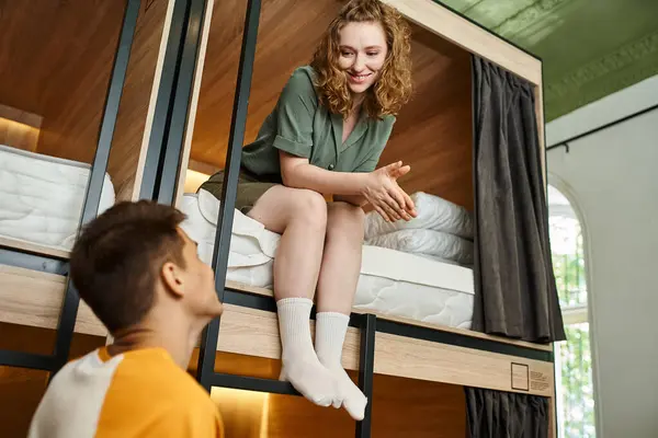 Joyeuse jeune femme assise sur un lit à deux étages et regardant petit ami dans une chambre confortable auberge — Photo de stock