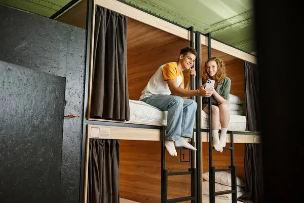 Joyeuse jeune femme montrant smartphone au petit ami sur les lits à deux étages dans la chambre confortable auberge — Photo de stock