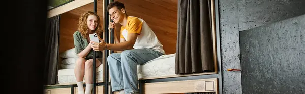 Femme jeune insouciante montrant smartphone au petit ami sur les lits à deux étages dans la chambre de l'auberge, bannière — Photo de stock