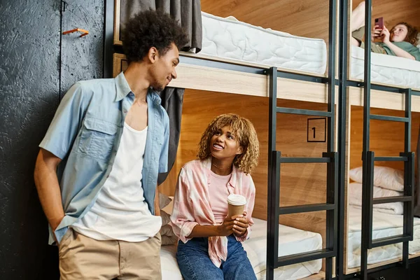 Smiling african american woman with paper cup talking to friend in comfortable room of youth hostel — Stock Photo