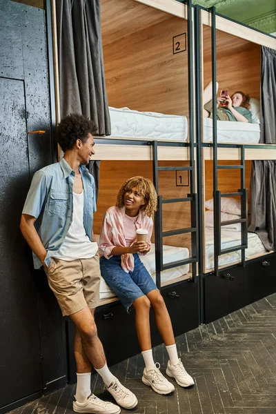 Heureuse femme afro-américaine avec tasse en papier parler à un ami dans la chambre de l'auberge avec lits à deux étages — Photo de stock