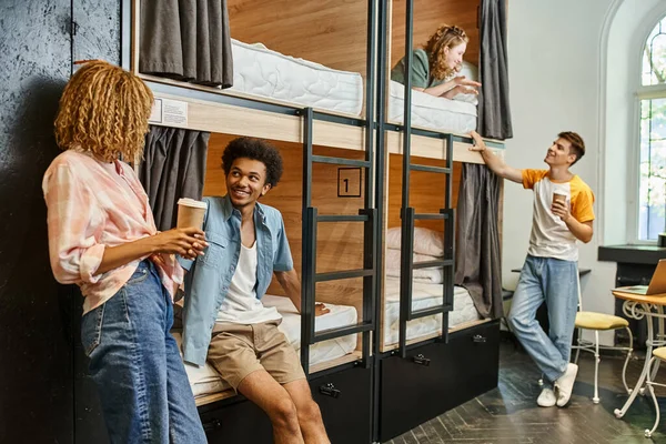 Cheerful multicultural roommates with coffee to go talking near double-decker beds in youth hostel — Stock Photo