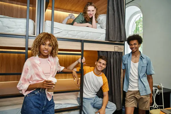 Alegres amigos multiétnicos mirando a la cámara cerca de camas de dos pisos en la acogedora habitación del albergue de estudiantes - foto de stock