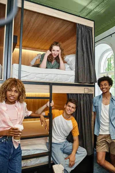 Copains multiethniques insouciants regardant la caméra près de lits à deux étages dans la chambre des étudiants auberge — Photo de stock
