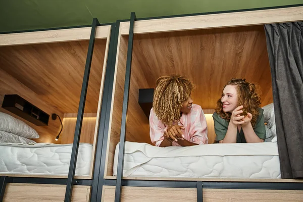 Carefree multicultural girlfriends lying and talking on double-decker bed in modern students hostel — Stock Photo