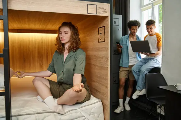 Young woman meditating on double-decker bed near friends with laptop in cozy room of students hostel — Stock Photo