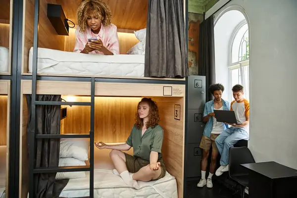 Youthful woman meditating on double-decker bed near multiethnic roommates with gadgets in hostel — Stock Photo