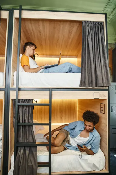 Estudiantes multiculturales que utilizan gadgets en camas de dos pisos en albergue juvenil contemporáneo, viajeros - foto de stock