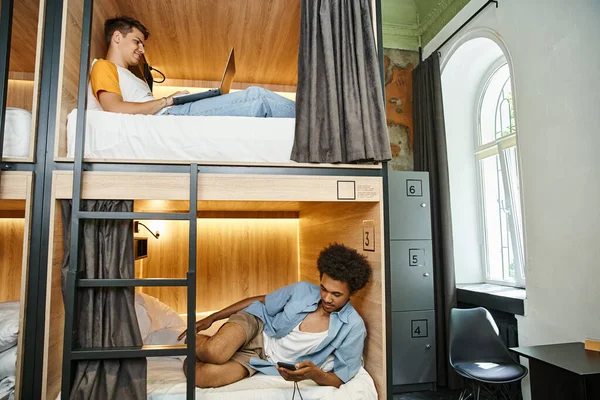 Amis interracial avec ordinateur portable et smartphone sur des lits à deux étages dans une auberge moderne, vacances — Photo de stock