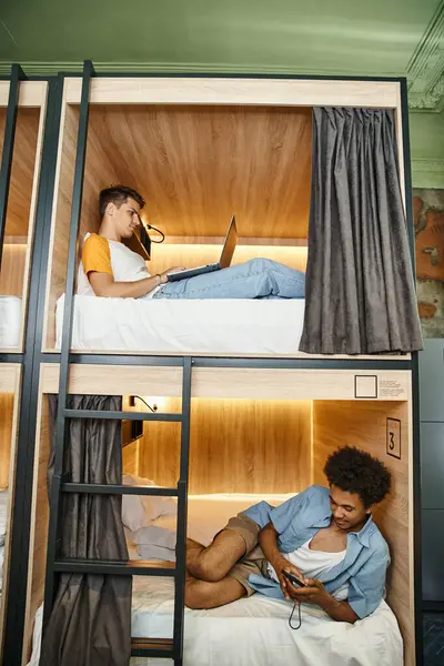 Étudiants multiethniques utilisant un ordinateur portable et un smartphone sur des lits à deux étages dans une auberge de jeunesse confortable — Photo de stock