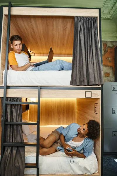Copains multiethniques joyeux avec ordinateur portable et smartphone se regardant sur les lits à deux étages — Photo de stock