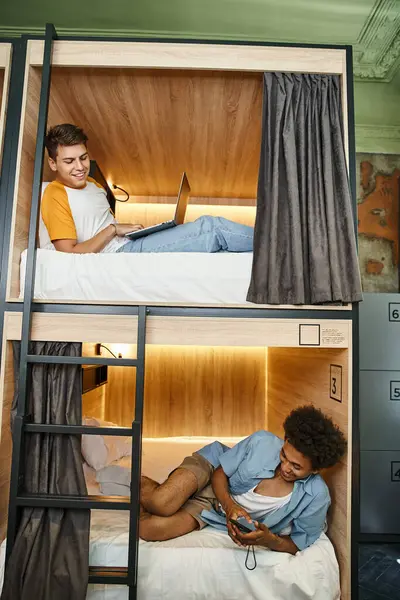 Joyful multiethnic roommates lying with laptop and smartphone on double-deckers beds in hostel — Stock Photo