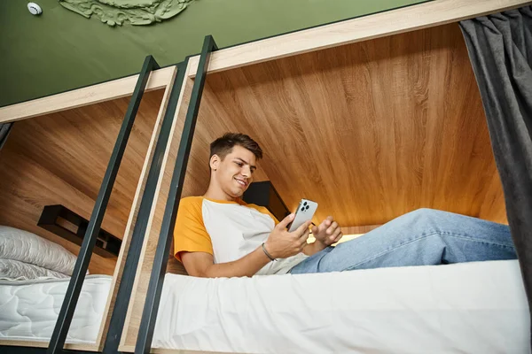 Vista de ángulo bajo de hombre joven navegar por Internet en el teléfono inteligente en la cama de dos pisos en el albergue - foto de stock