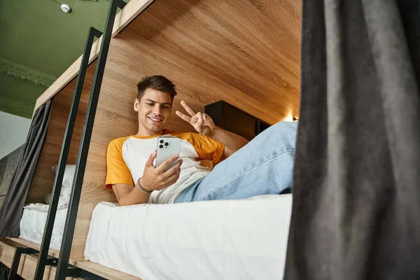 Lächelnder Student zeigt Siegeszeichen bei Videoanruf auf Smartphone auf Doppelstockbett in Hostel — Stockfoto
