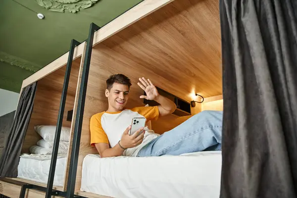 Alegre estudiante saludando la mano durante la videollamada en el teléfono inteligente en la cama de dos pisos en el albergue - foto de stock