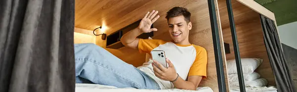 Alegre chico saludando la mano durante la videollamada en cama de dos pisos en acogedor albergue de estudiantes, pancarta - foto de stock