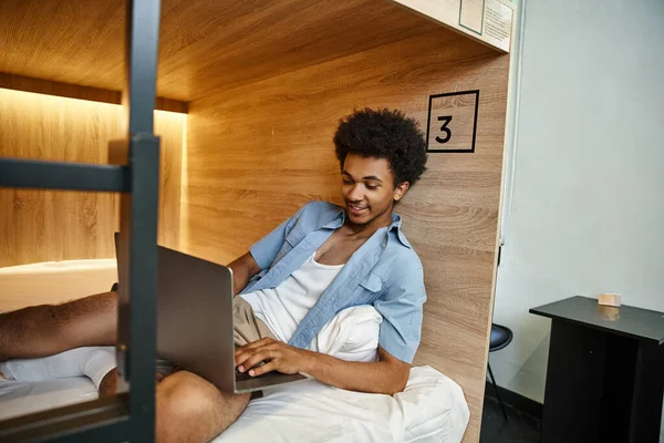 Heureux et élégant réseau étudiant afro-américain sur ordinateur portable sur lit double étage en auberge — Photo de stock