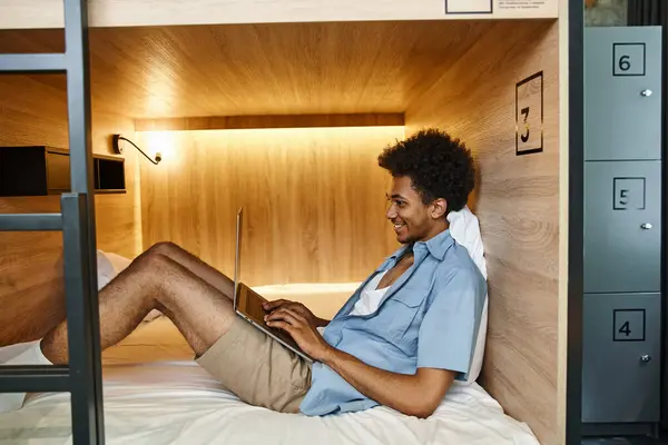 Vista lateral do homem americano africano alegre digitando no laptop na cama acolhedora de dois andares no albergue — Fotografia de Stock
