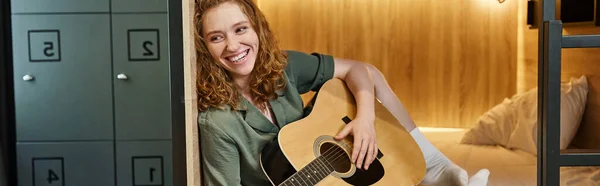 Joyful young woman playing acoustic guitar on double-decker bed in comfortable hostel room, banner — Stock Photo