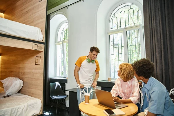 Young student looking at african american roommates near laptop in cozy hostel room, travelers — Stock Photo