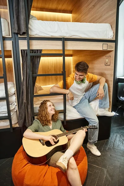 Smiling woman playing acoustic guitar to boyfriend sitting on double-decker bed in hostel room — Stock Photo