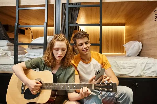 Jeune femme jouant de la guitare acoustique au petit ami avec smartphone dans la chambre confortable de l'auberge des étudiants — Photo de stock