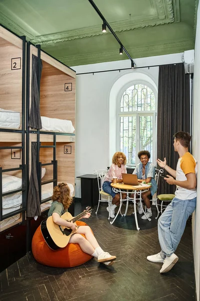 Multiethnic friends with laptop and guitar talking in cozy room of modern students hostel, leisure — Stock Photo