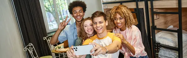 Cheerful young man taking selfie on smartphone with trendy multicultural friends in hostel, banner — Stock Photo