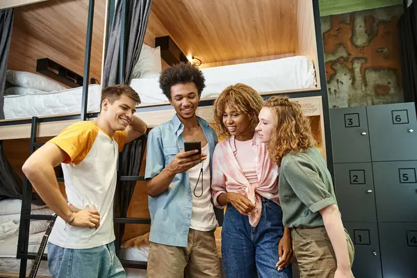 Smiling african american man browsing social media on smartphone near interracial friends in hostel — Stock Photo