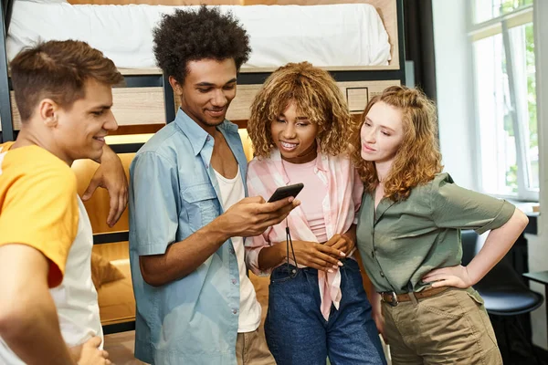 Homme afro-américain positif avec téléphone portable près de colocataires multiculturels dans l'auberge des étudiants — Photo de stock