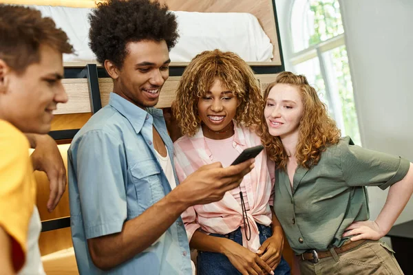 Pleased african american man browsing internet on smartphone near interracial mates in youth hostel — Stock Photo