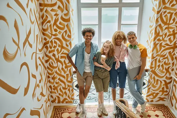 Беззаботные стильные мультикультурные друзья смотрят на камеру на лестнице с окрашенными стенами в общежитии — стоковое фото