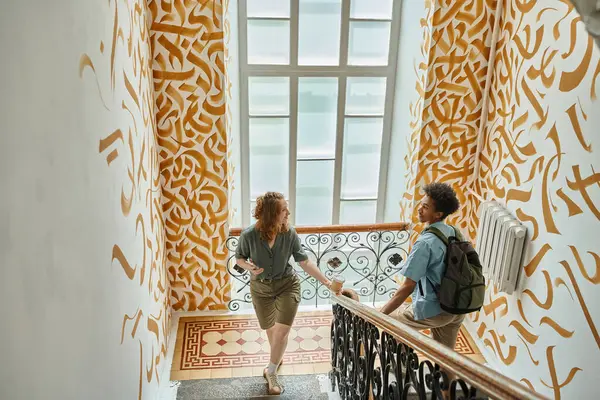 Щаслива жінка і афроамериканський студент ходить по сходах з барвистими стінами в гуртожитку — стокове фото