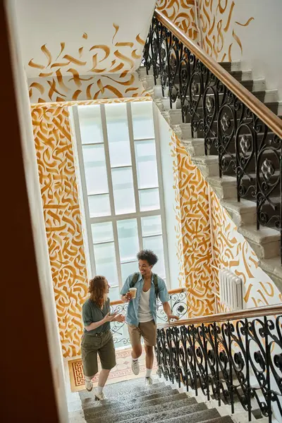 Feliz casal multiétnico andando e falando em escadaria com paredes coloridas no albergue moderno — Fotografia de Stock