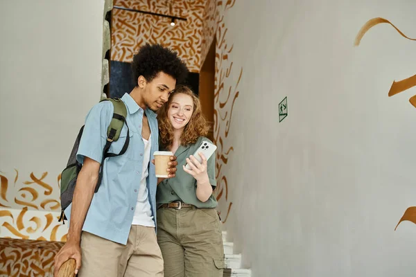 Joyful interracial couple with smartphone and coffee to go walking on staircase of students hostel — Stock Photo