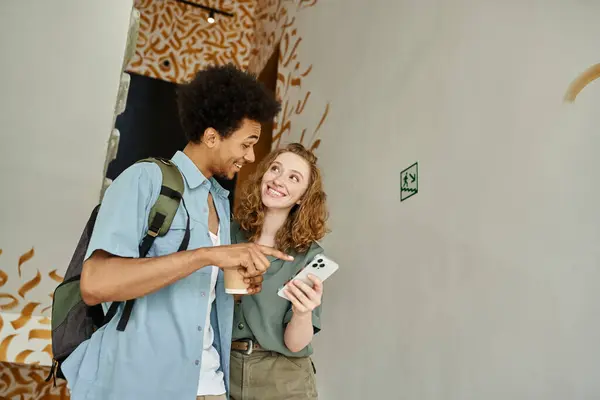 Веселый африканский американец указывает на смартфон рядом с молодой девушкой на лестнице общежития — стоковое фото