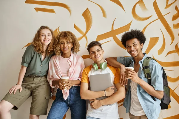 Cheerful multiethnic students with coffee to go and laptop smiling at camera on staircase of hostel — Stock Photo