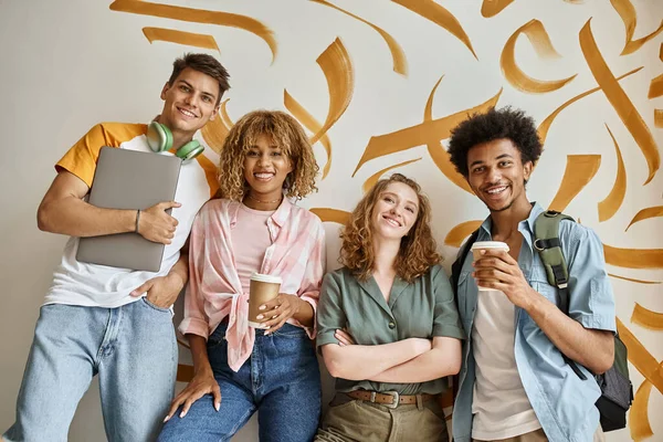 Carefree multiethnic friends with coffee to go and laptop smiling at camera on staircase of hostel — Stock Photo