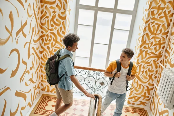 Young cheerful multiethnic students with backpacks walking on staircase of modern hostel — Stock Photo