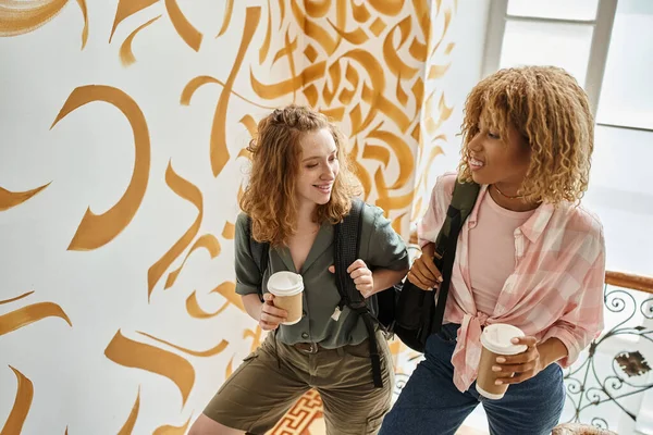 Cheerful interracial female friends with paper cups and backpacks on staircase of modern hostel — Stock Photo