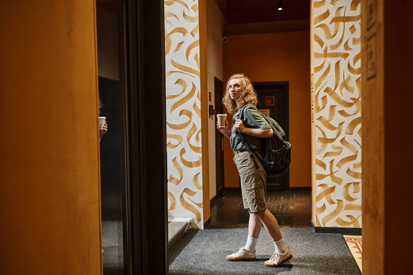 Mujer joven con taza de papel de pie en el pasillo con paredes de colores en el albergue de estudiantes, viajero - foto de stock