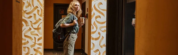 Estudiante femenina con mochila y taza de papel en el pasillo de albergue juvenil contemporáneo, pancarta - foto de stock