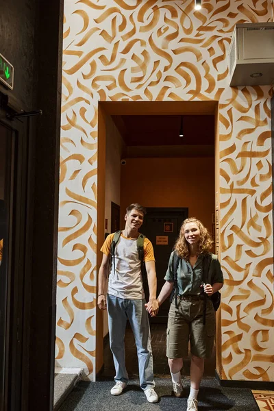 Feliz pareja joven cogidos de la mano y mirando a la cámara en el pasillo del albergue con diseño moderno - foto de stock