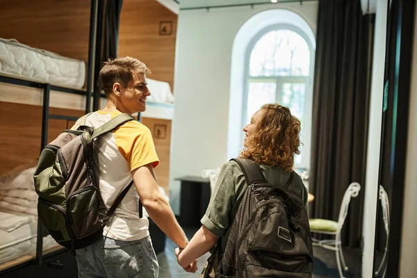 Happy young couple with backpacks holding hands and looking at each other in cozy room of hostel — Stock Photo