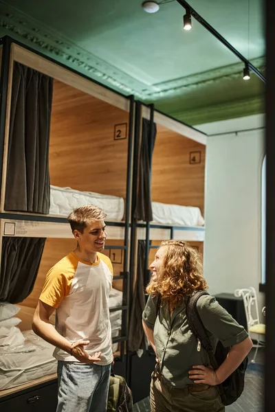 Cheerful and young couple with backpacks talking in cozy room of students hotel, travelers — Stock Photo