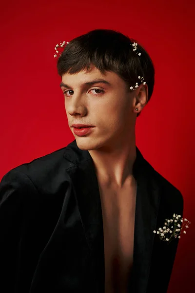 Красивый молодой человек в стильном наряде с цветами в волосах, смотрящий в камеру на красном фоне — стоковое фото